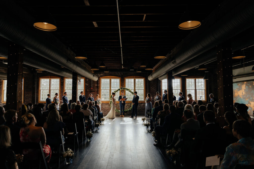 A wedding ceremony in the Tinker House event space, seen from the back of the room, with tall arched windows and guests seated on both sides of the aisle