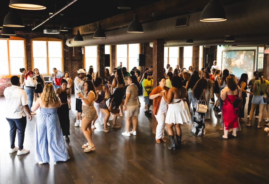 A packed dance floor at a daytime community party in the Tinker House event space, with sunlight through the tall windows