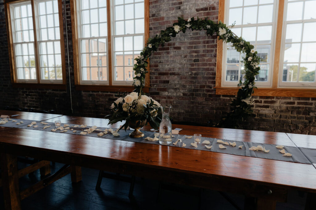 A long head table set with candles and white flowers under a greenery wreath in front of the brick wall and arched windows at Tinker House