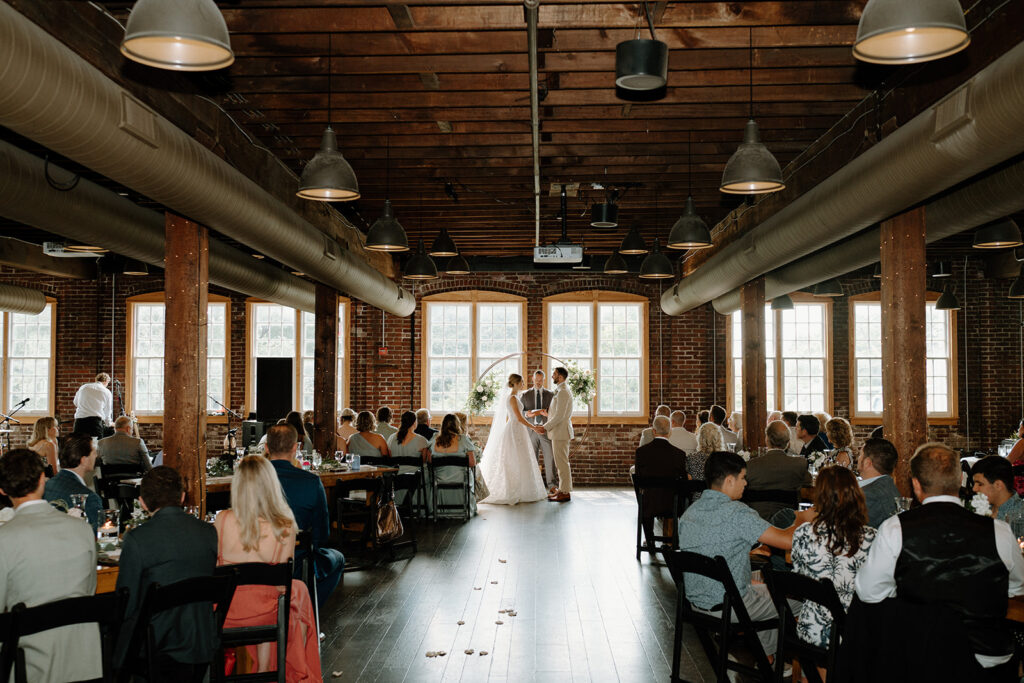 Wedding guests at dinner in the Tinker House event space, with timber beams, Edison pendants and a wall of tall factory windows